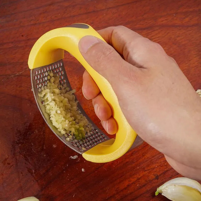 Stainless steel manual garlic press with coloured non-slip handle on a kitchen counter.
