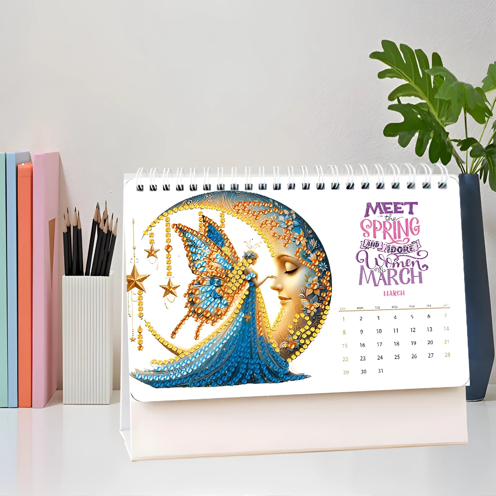 Frameless white card desk calendar with cartoon diamond painting design and stick-on crystals on a desk surface.