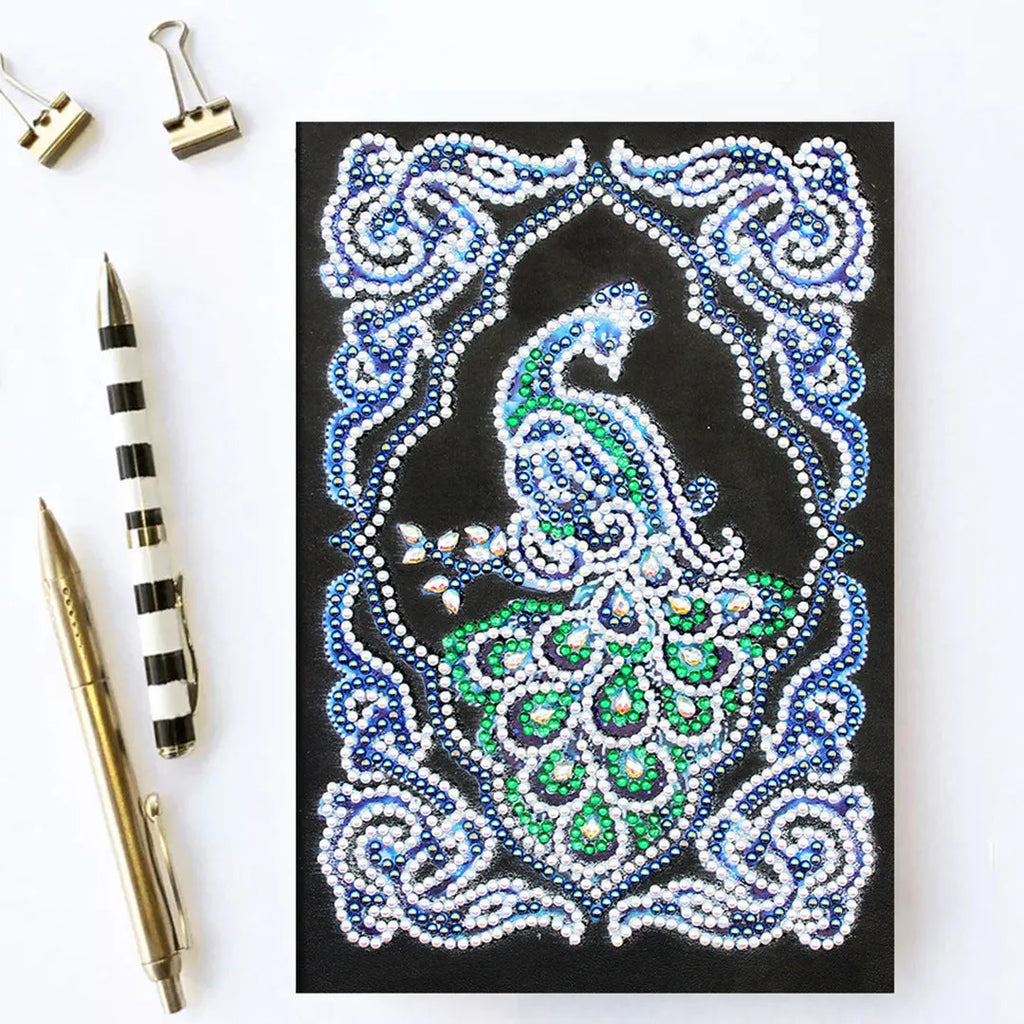 A5 notebook with a sparkling peacock diamond painting on the cover, shown next to open lined pages.