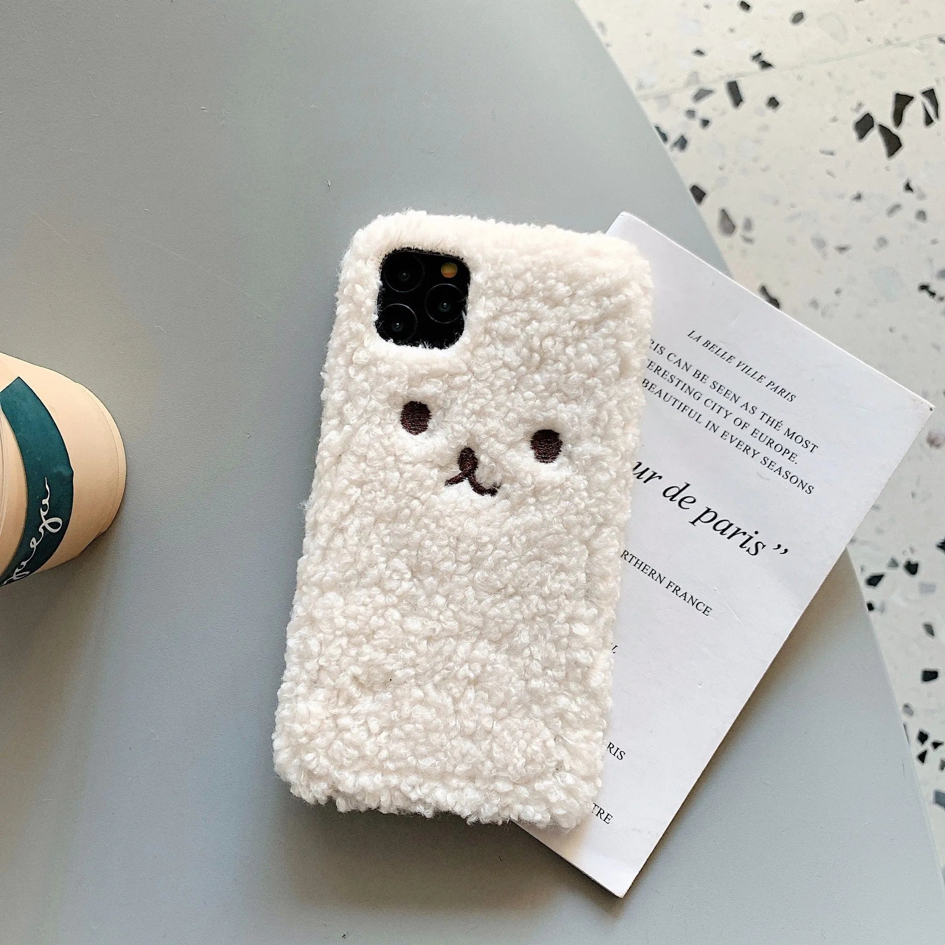 Brown embroidered plush bear face on a clear TPU phone case for iPhone, showing precise button and camera cutouts.