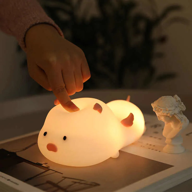 Blue rechargeable pig-shaped silicone night light with soft glow on a bedside table.