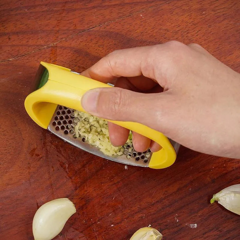 Stainless steel manual garlic press with coloured non-slip handle on a kitchen counter.