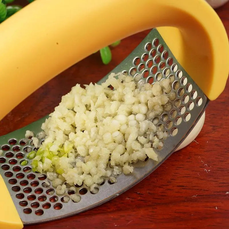 Stainless steel manual garlic press with coloured non-slip handle on a kitchen counter.