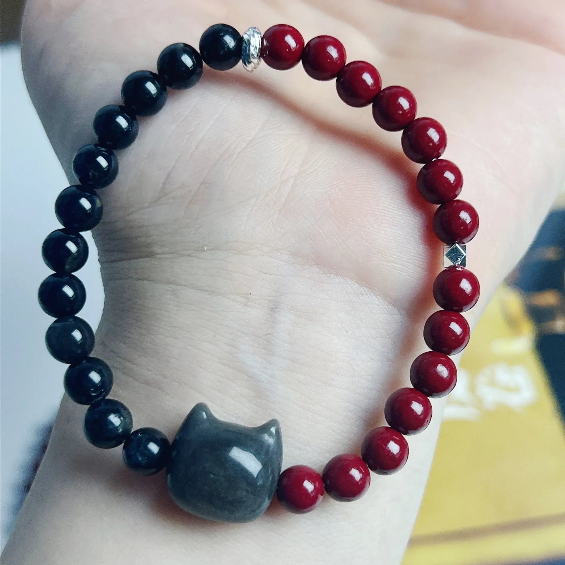 Handmade obsidian and glass bead bracelet with a cat charm and red string, showing adjustable stainless steel clasp.
