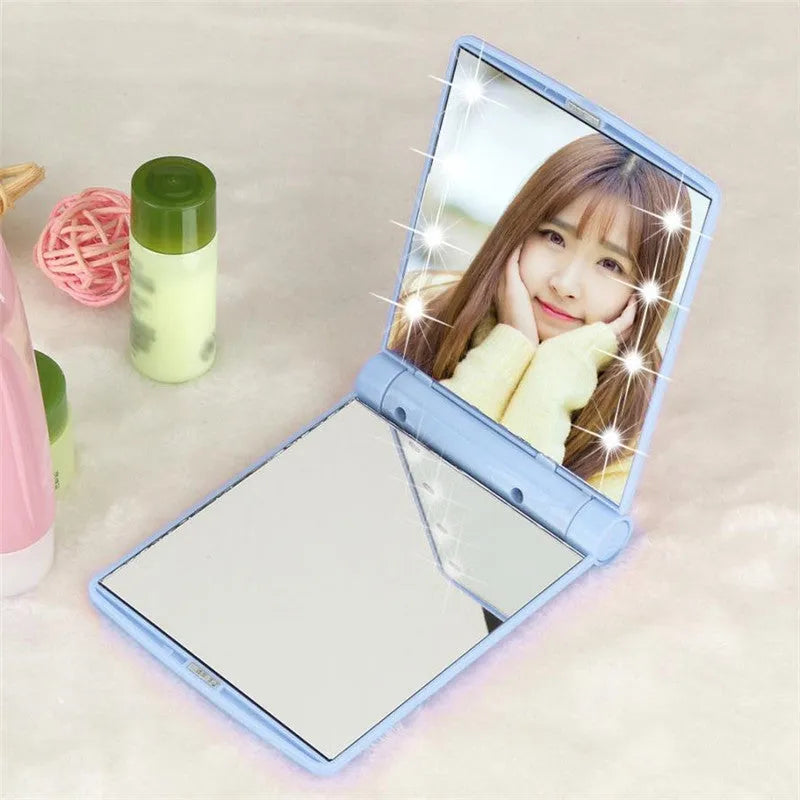 Folding LED pocket cosmetic mirror in black, blue, pink, or white with protective lid and compact plastic casing for handbag or travel use.