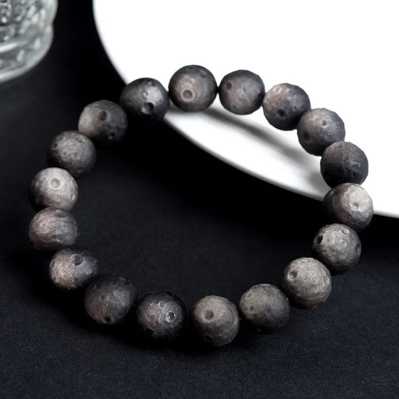 A natural obsidian crater ball bracelet in gold or silver meteorite colour on a wrist, showing textured bead detail.