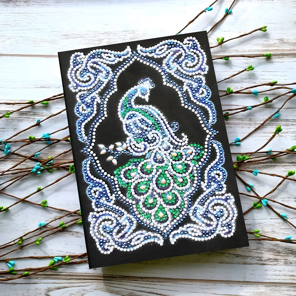 A5 notebook with a sparkling peacock diamond painting on the cover, shown next to open lined pages.