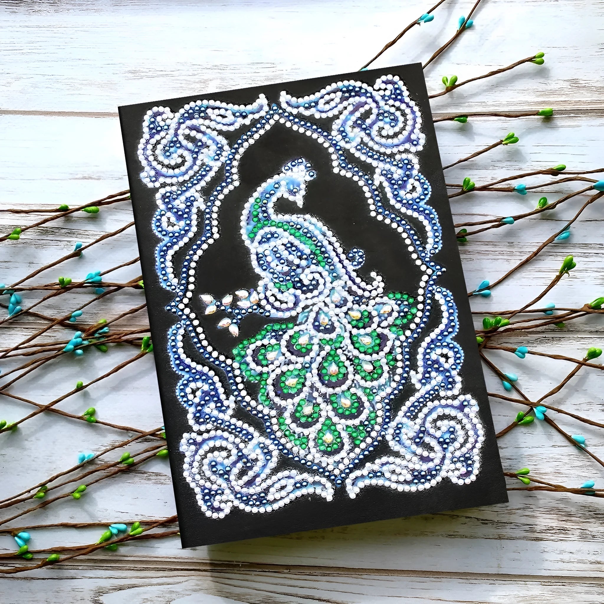A5 notebook with a sparkling peacock diamond painting on the cover, shown next to open lined pages.