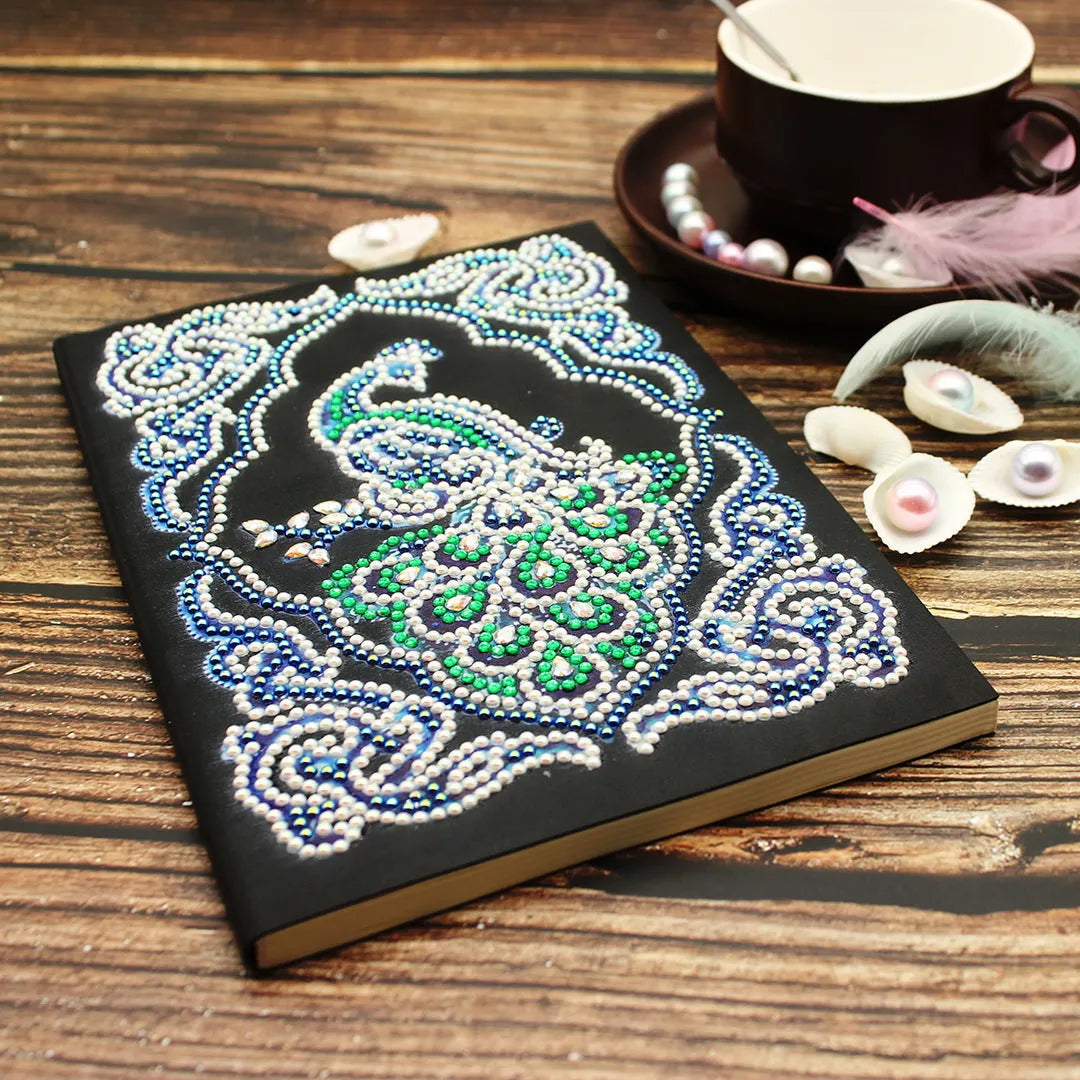 A5 notebook with a sparkling peacock diamond painting on the cover, shown next to open lined pages.