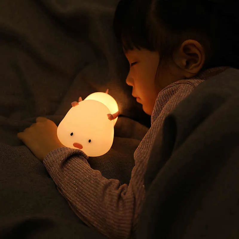 Blue rechargeable pig-shaped silicone night light with soft glow on a bedside table.