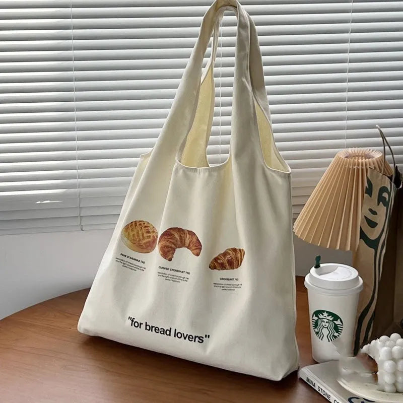 Beige canvas shoulder bag with croissant and bread print showing front panel design and shoulder strap