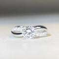 Silver ring with a 1 karat moissanite stone in a twisted four-claw setting, showing geometric lines and a cross design.
