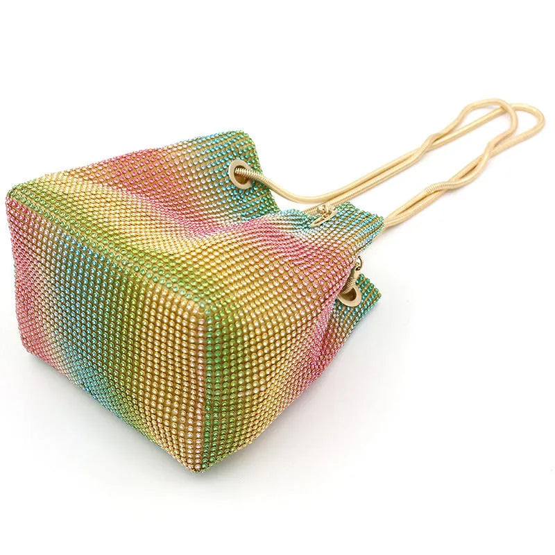 Soft rainbow iridescent bucket bag covered in sparkling rhinestones with a shoulder strap on a white background.