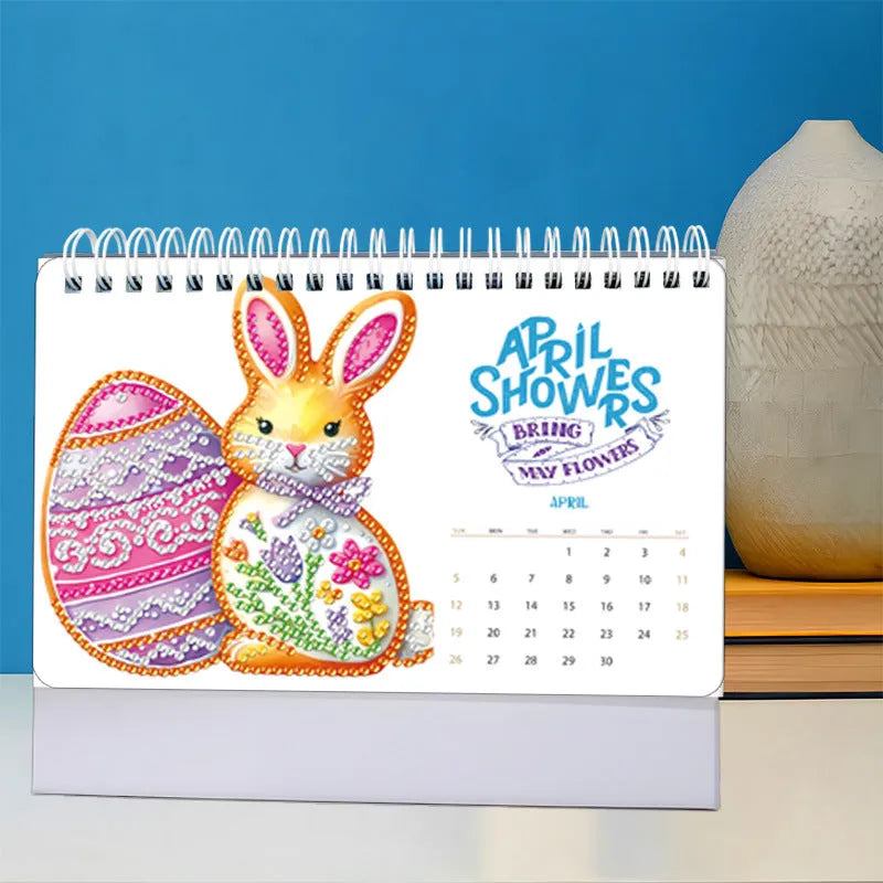 Frameless white card desk calendar with cartoon diamond painting design and stick-on crystals on a desk surface.