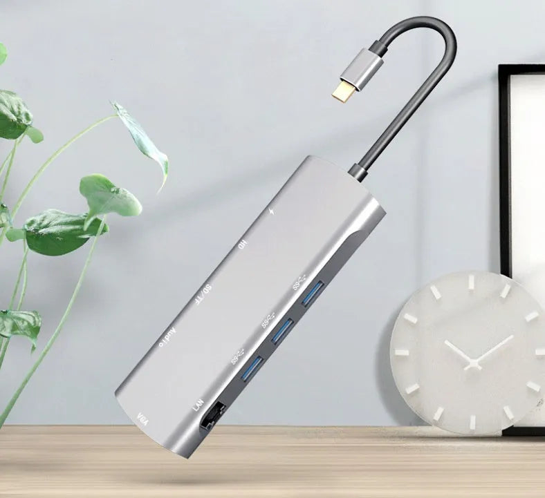 Grey aluminium USB-C docking station with HDMI, VGA, USB 3.0, and PD charge ports on a wooden desk.