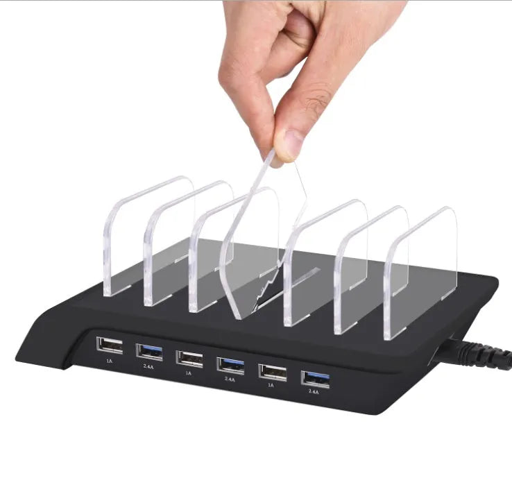 Six-port USB mobile phone charging stand in silver with multiple phones and tablets inserted, on a wooden desk