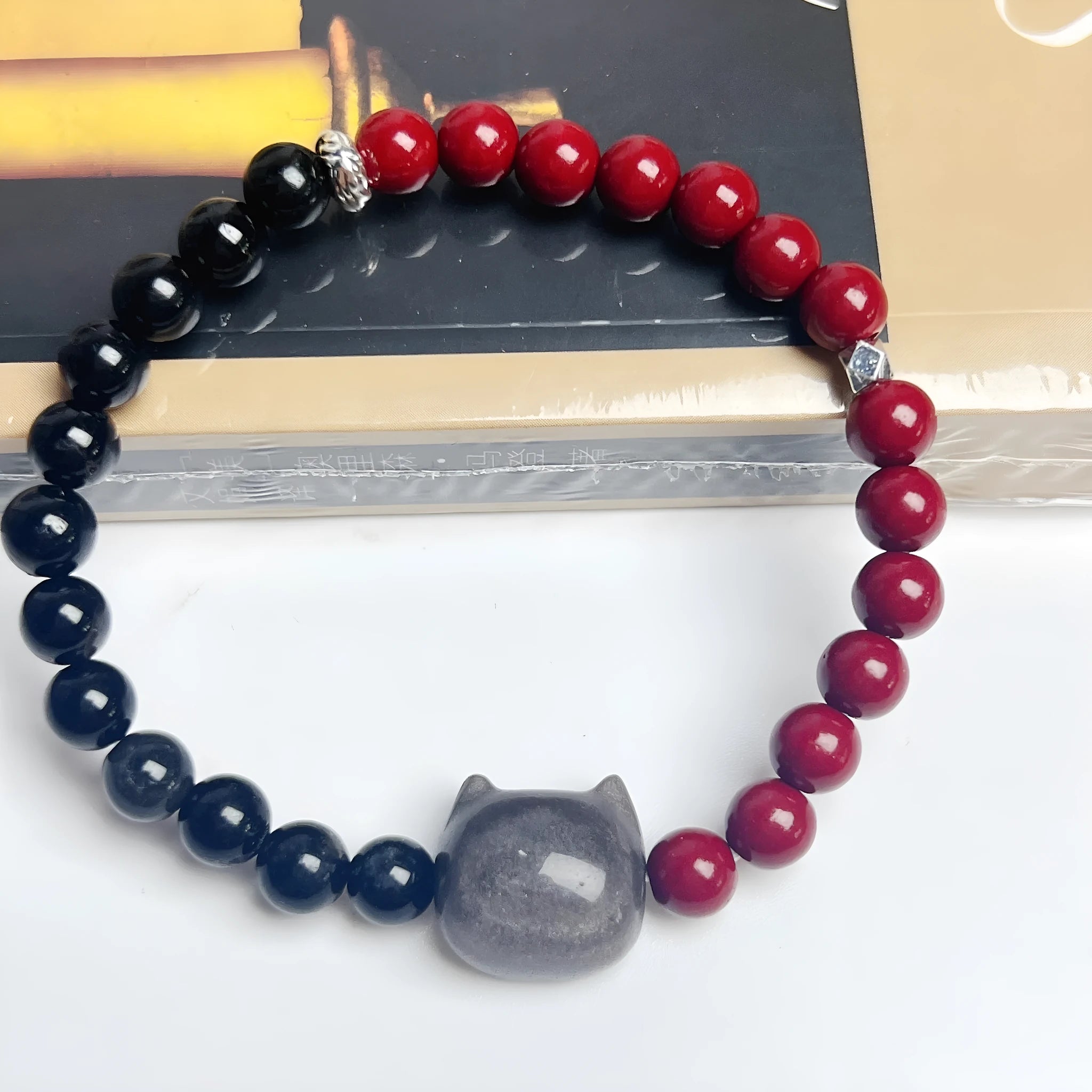 Handmade obsidian and glass bead bracelet with a cat charm and red string, showing adjustable stainless steel clasp.