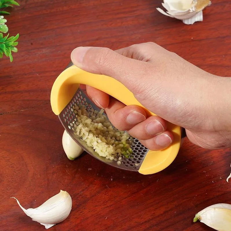 Stainless steel manual garlic press with coloured non-slip handle on a kitchen counter.