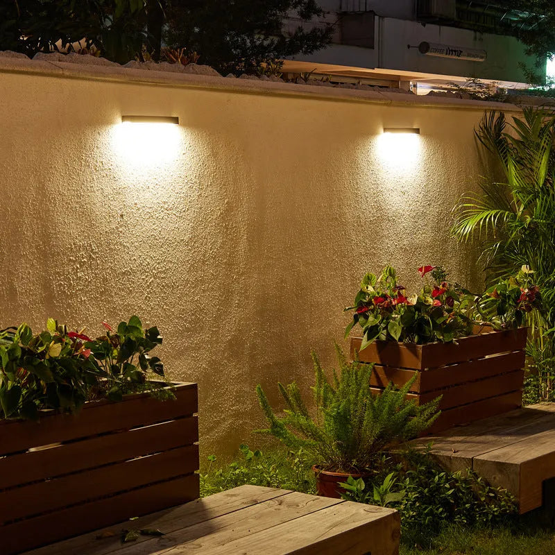 Black solar-powered outdoor wall lamp with integrated solar panel, mounted on a garden wall.