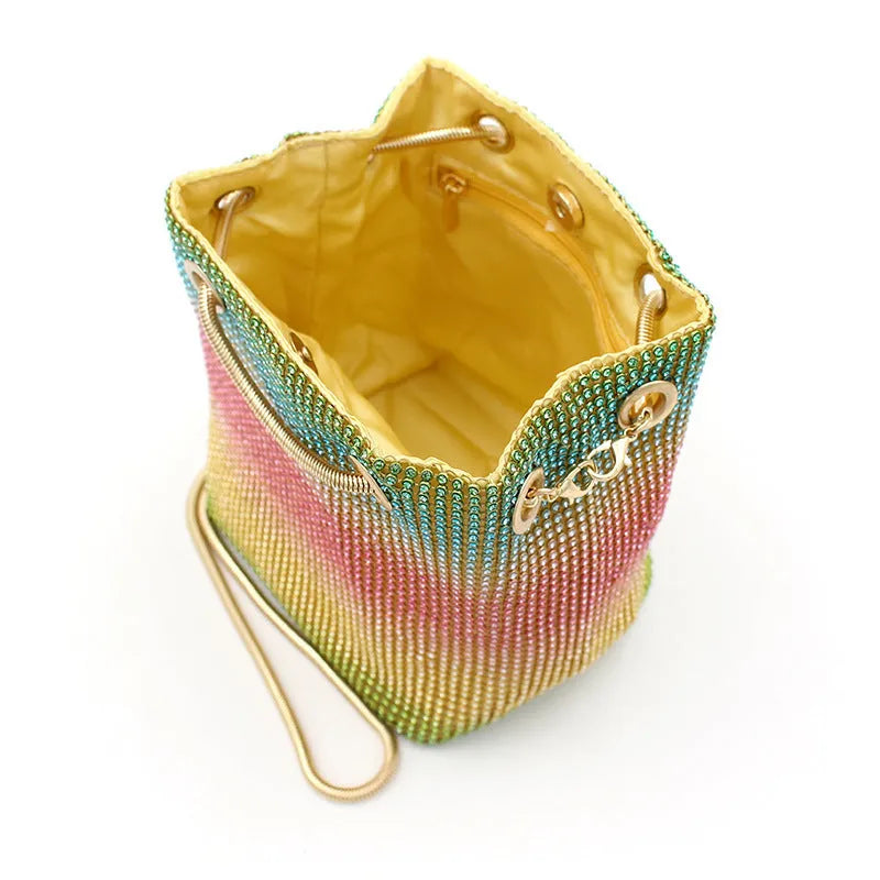 Soft rainbow iridescent bucket bag covered in sparkling rhinestones with a shoulder strap on a white background.