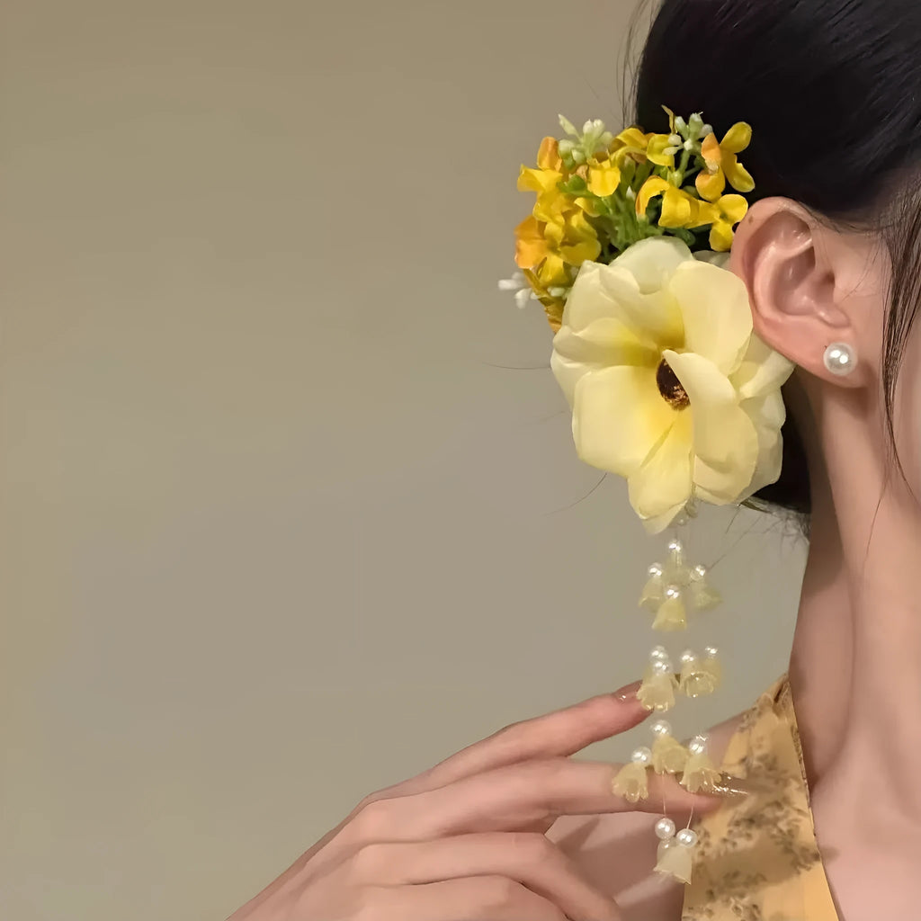 Close-up of a women's hair clip with a white pearl, yellow fabric flowers, and a long yellow tassel on a dark background.