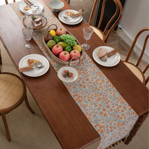 Beige jacquard table runner with floral pattern and long tassel fringes on a table.