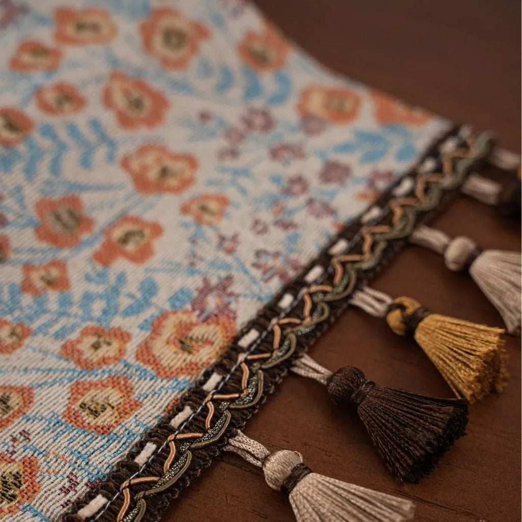 Beige jacquard table runner with woven leaf pattern and long knotted tassels draped on a wooden dining table.