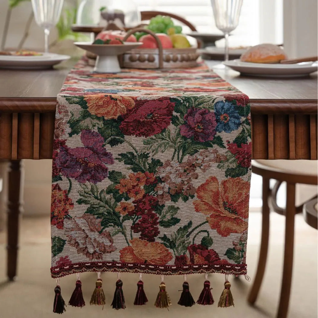 Beige jacquard table runner with woven leaf pattern and long knotted tassels draped on a wooden dining table.
