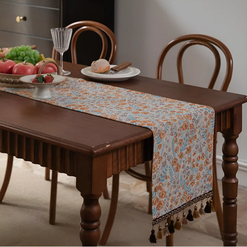 Beige jacquard table runner with floral pattern and long tassel fringes on a table.
