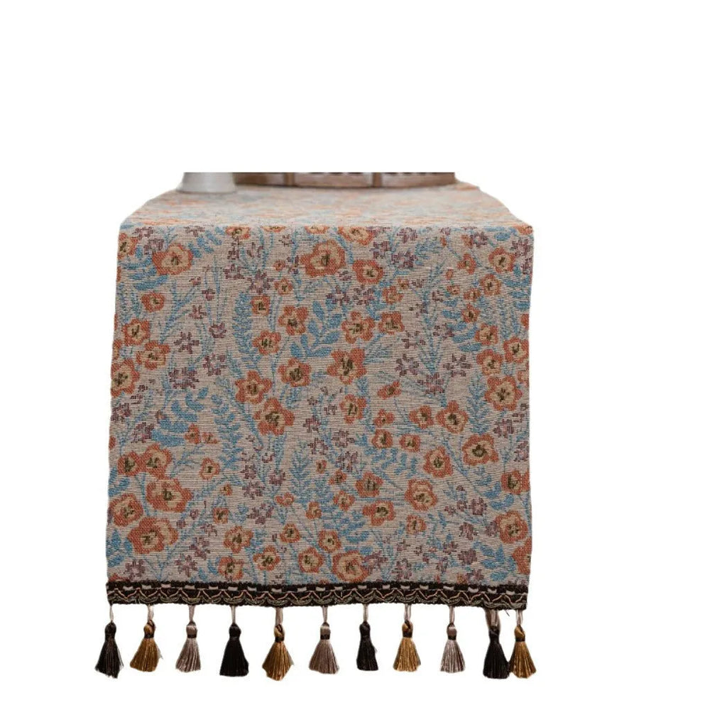 Beige jacquard table runner with woven leaf pattern and long knotted tassels draped on a wooden dining table.