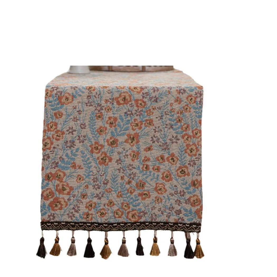 Beige jacquard table runner with woven leaf pattern and long knotted tassels draped on a wooden dining table.