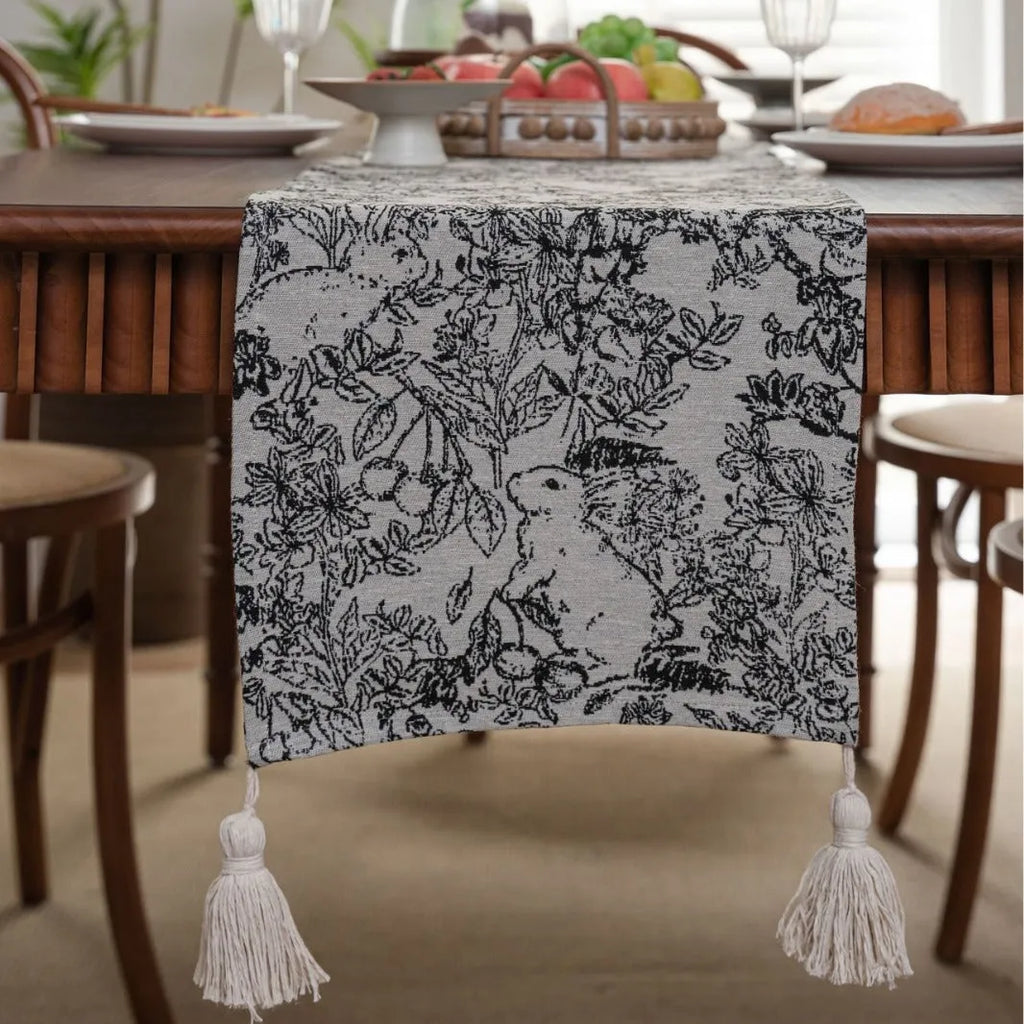 Beige jacquard table runner with woven leaf pattern and long knotted tassels draped on a wooden dining table.