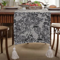 Beige jacquard table runner with woven leaf pattern and long knotted tassels draped on a wooden dining table.