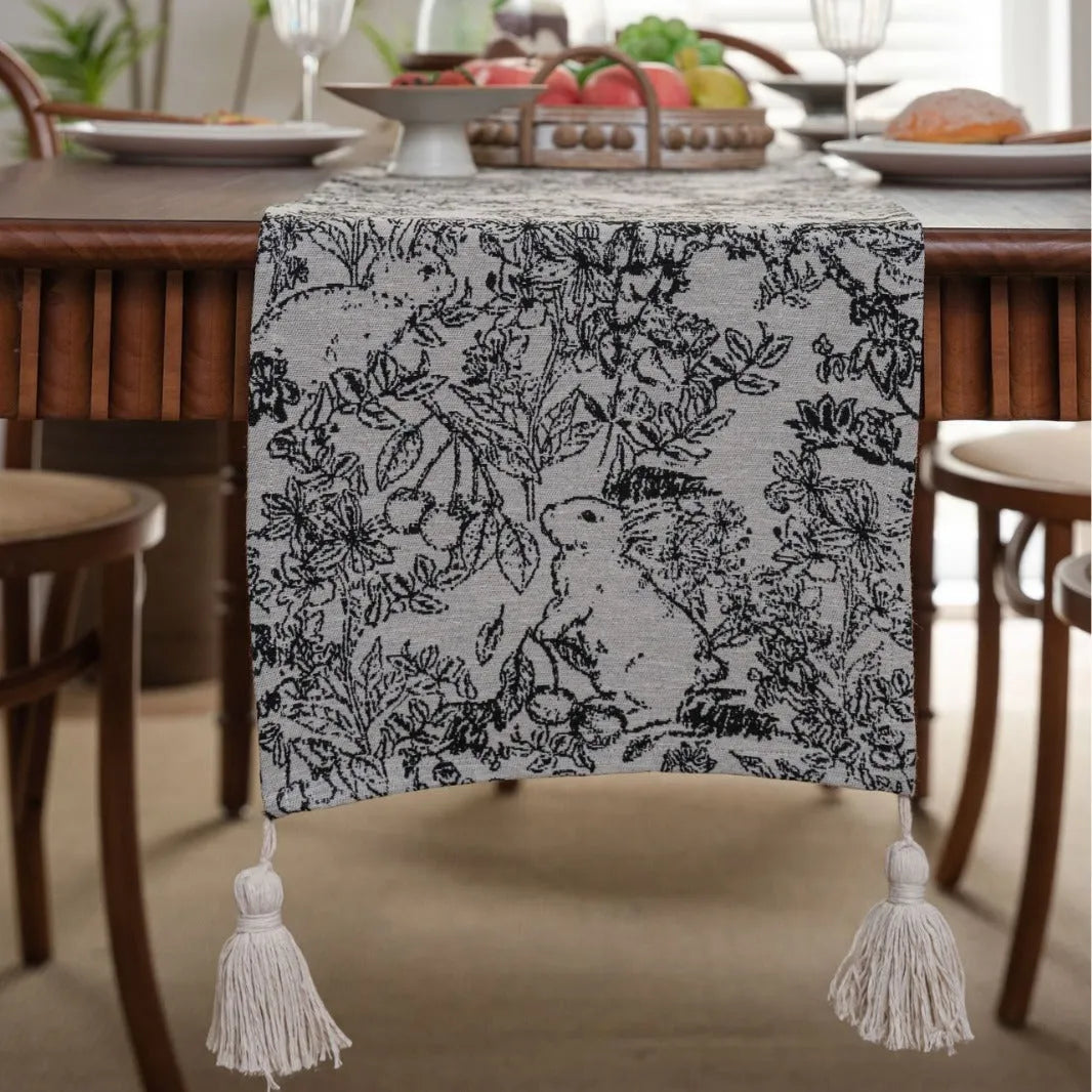 Beige jacquard table runner with woven leaf pattern and long knotted tassels draped on a wooden dining table.