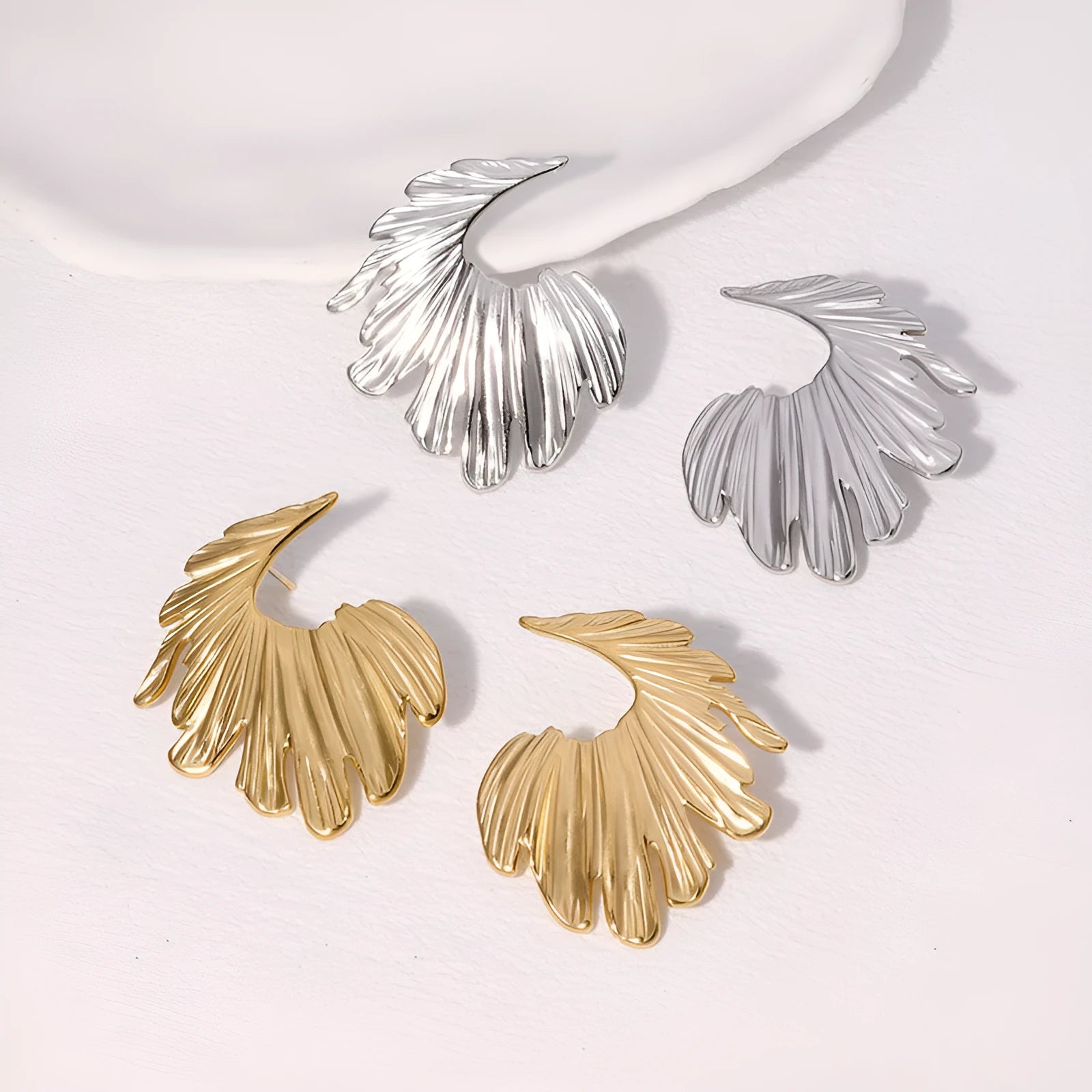 A pair of stainless steel stud earrings in a leaf shape, shown in gold colour and steel colour finishes.