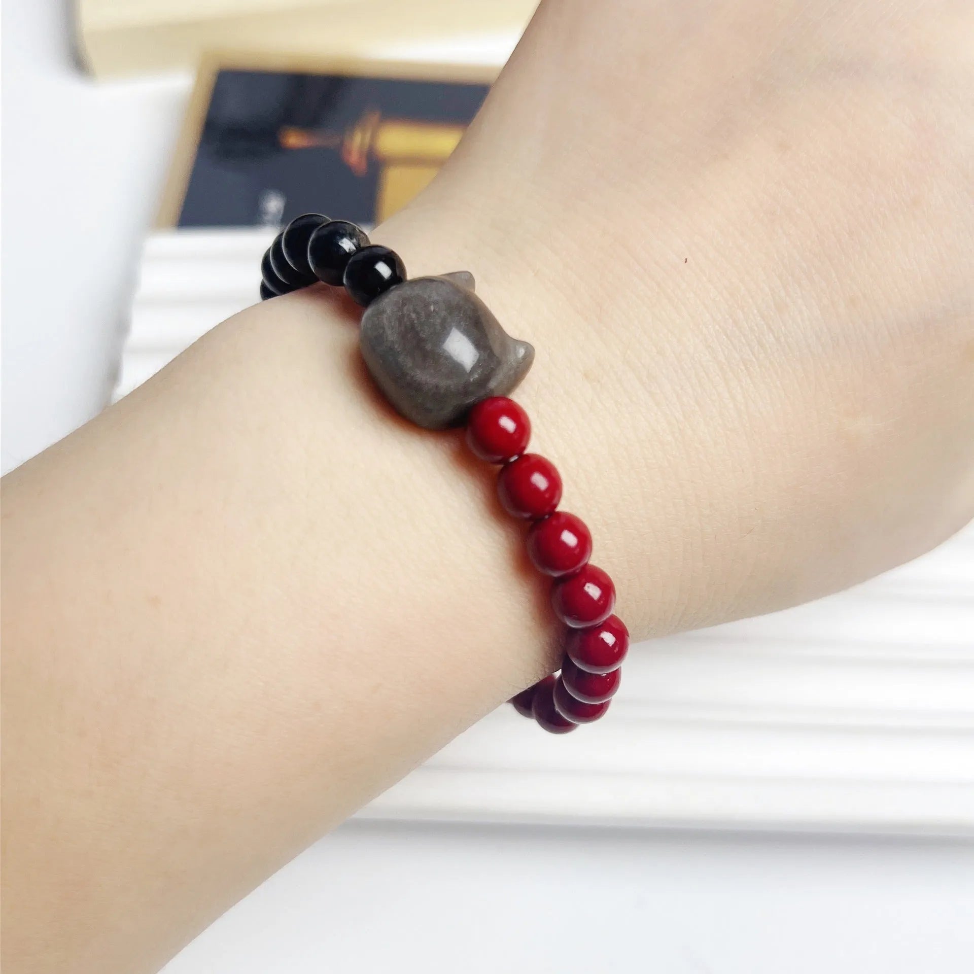 Handmade obsidian and glass bead bracelet with a cat charm and red string, showing adjustable stainless steel clasp.