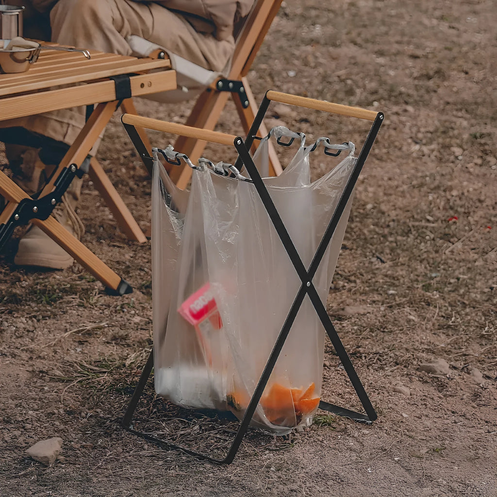 Outdoor black iron folding garbage storage hanger with wooden handle holding a bin bag.