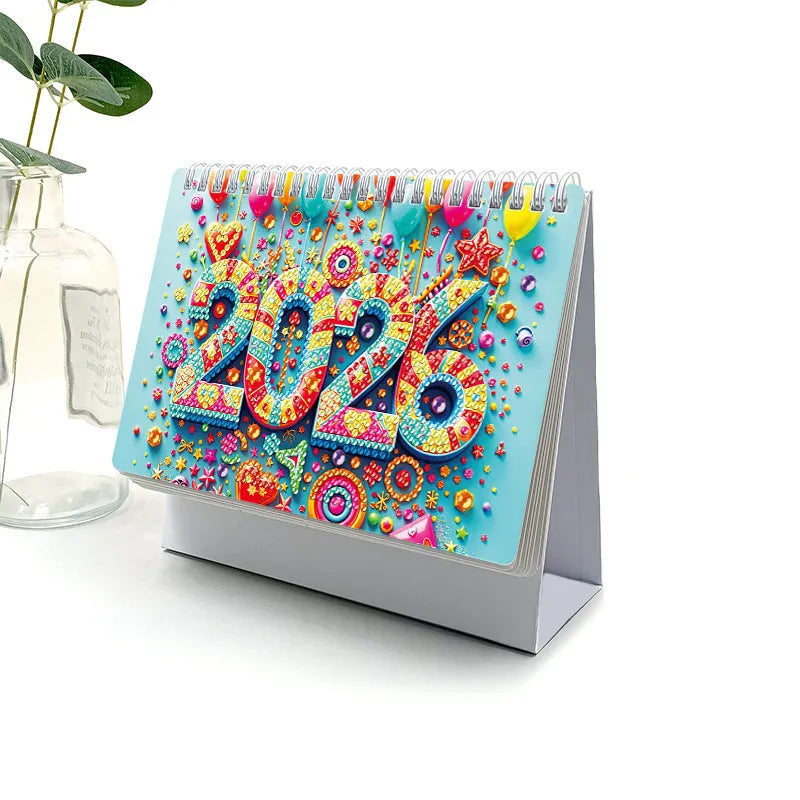 Frameless white card desk calendar with cartoon diamond painting design and stick-on crystals on a desk surface.